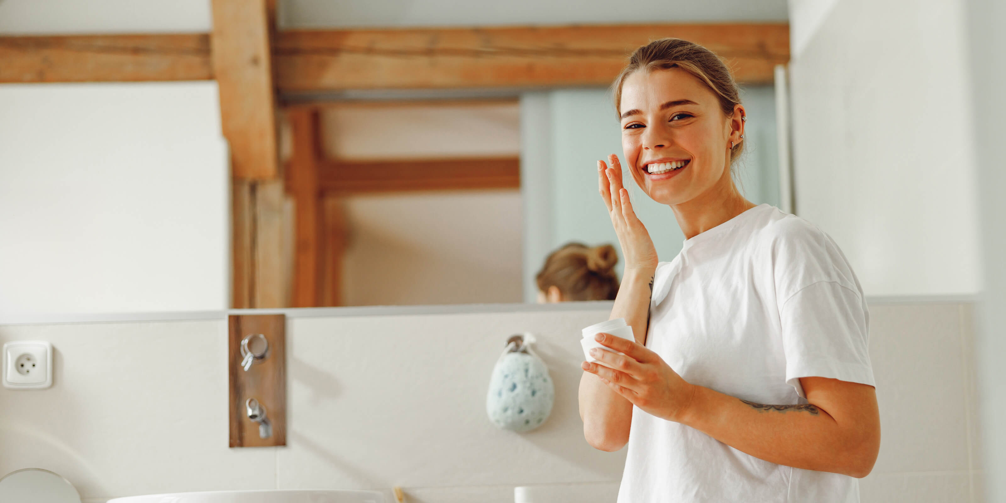 A girl smiling in front of a mirror putting skincare cream on her face.