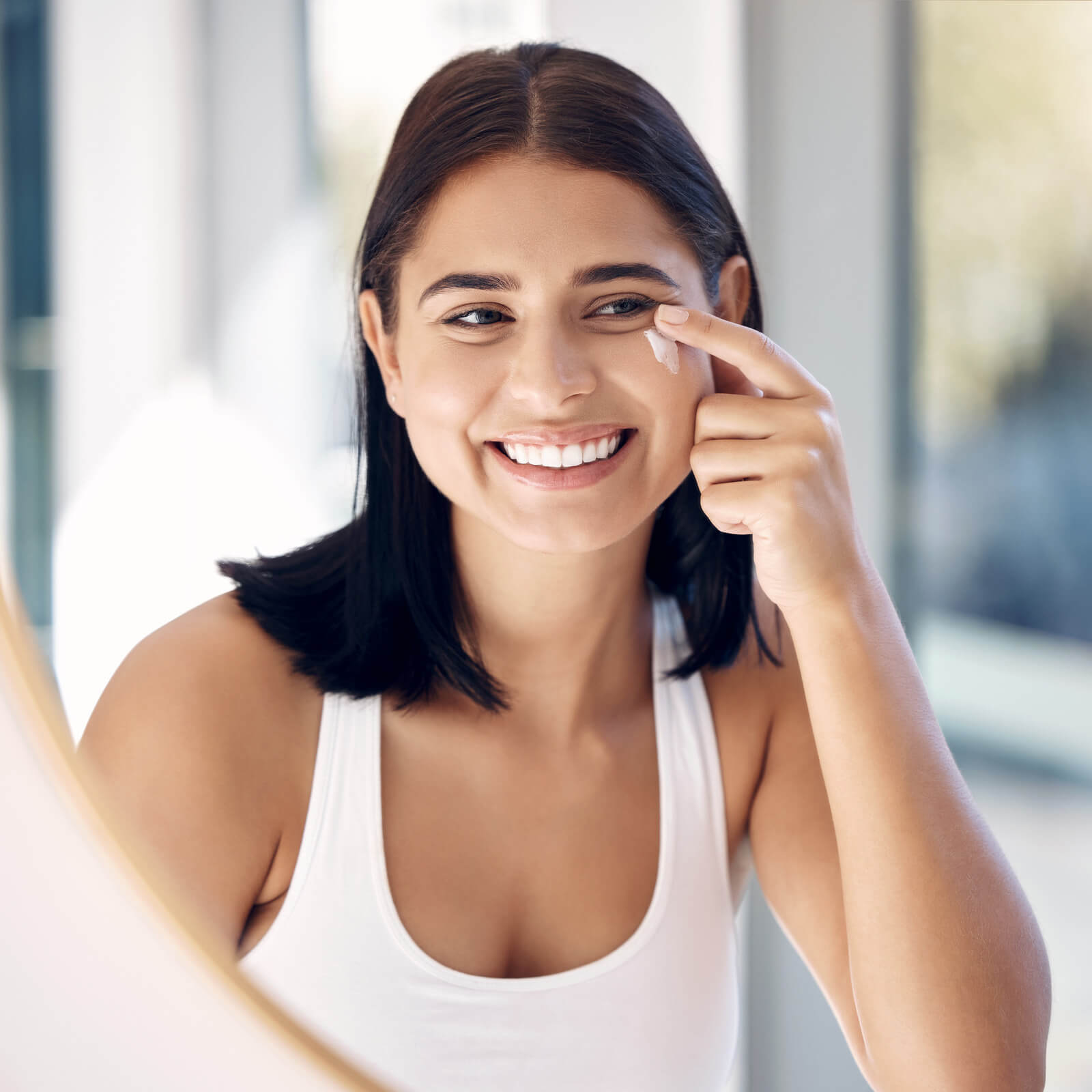 A woman smiling into a mirror putting cream under her right eye.