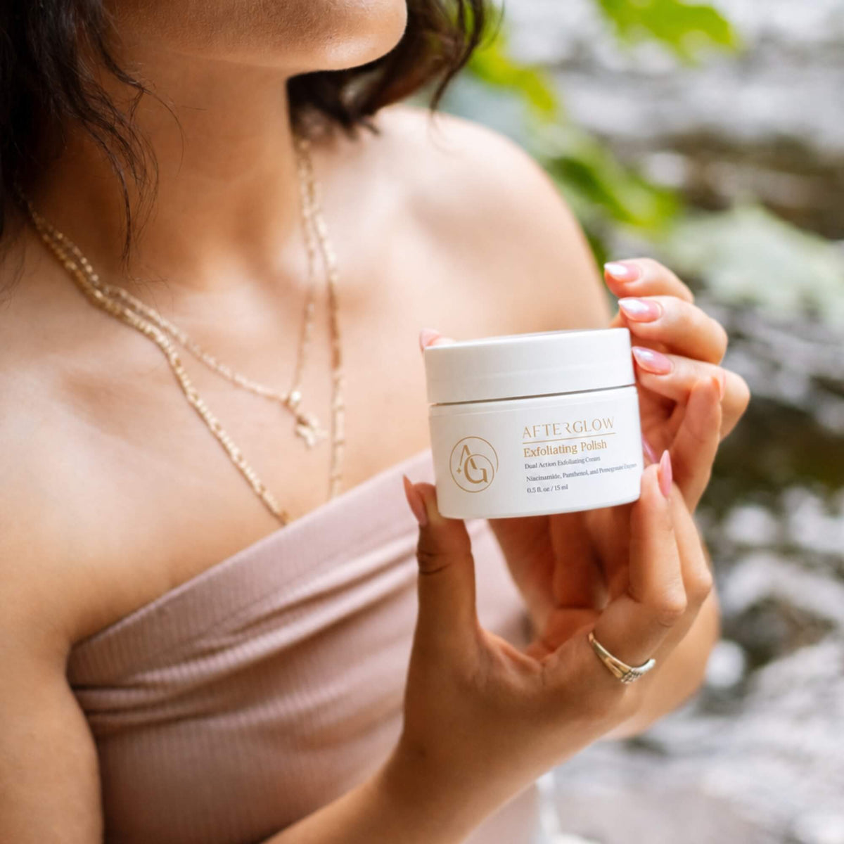 A woman in a strapless top holds a white jar of Exfoliating polish in front of a blurred outdoor background.